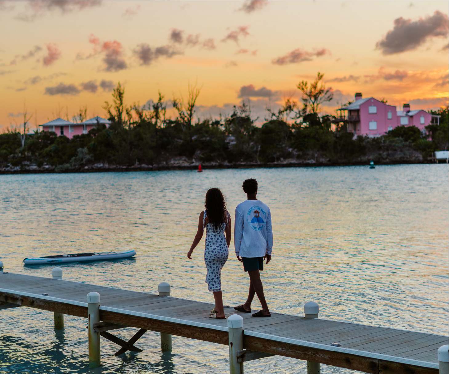 A couple walks hand in hand on a wooden dock by a calm body of water during sunset, with houses and trees on the opposite shore and colorful clouds in the sky.