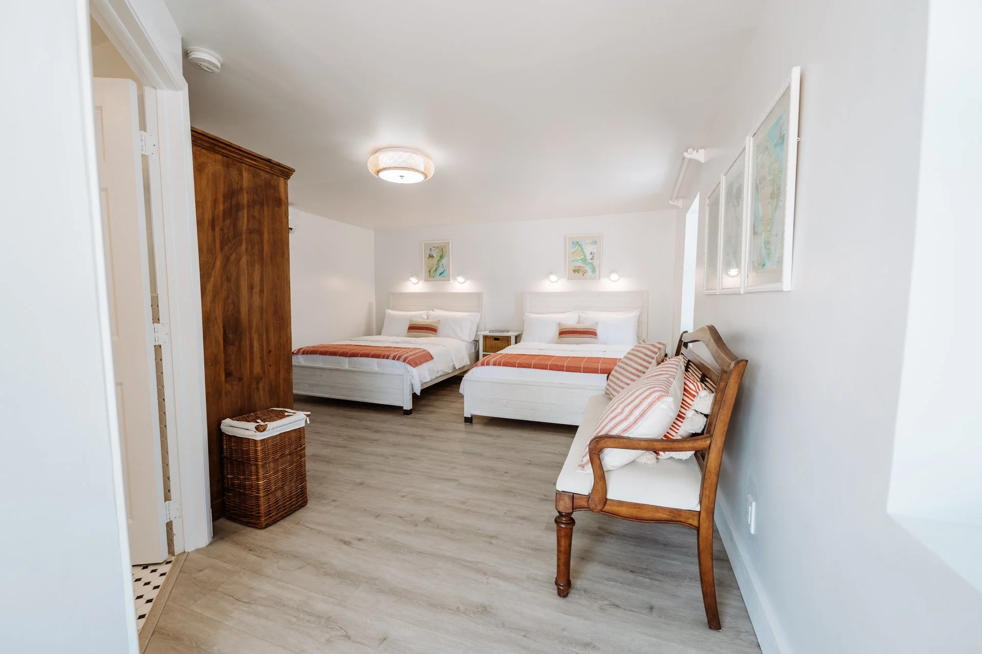 A bright hotel room with two white beds, orange striped bedding, a wooden wardrobe, a wicker laundry basket, a wooden bench with throw pillows, a wall-mounted art piece, and a ceiling light.