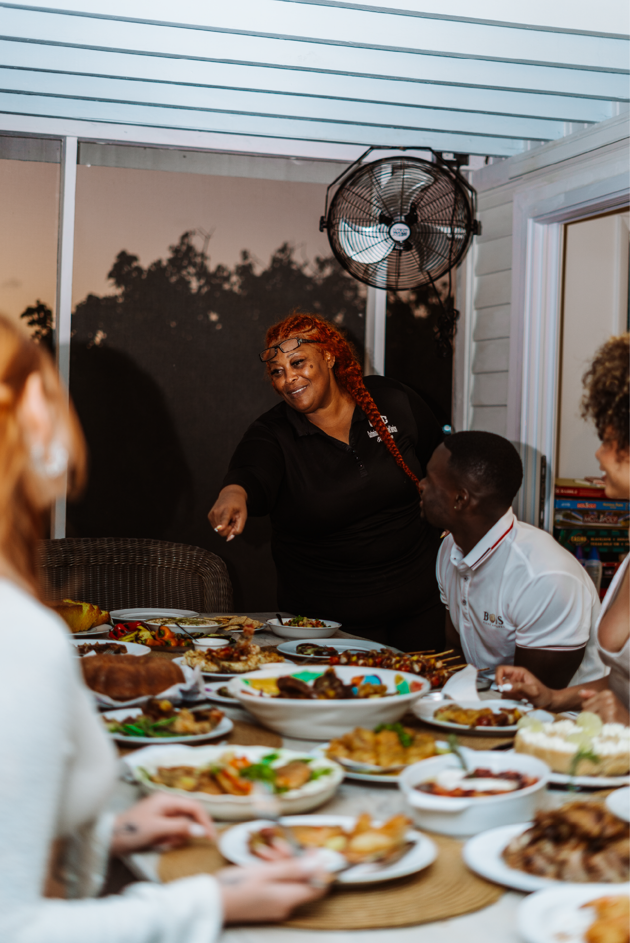 A woman with red hair and glasses pointing at a person while standing behind a table full of food during a gathering.