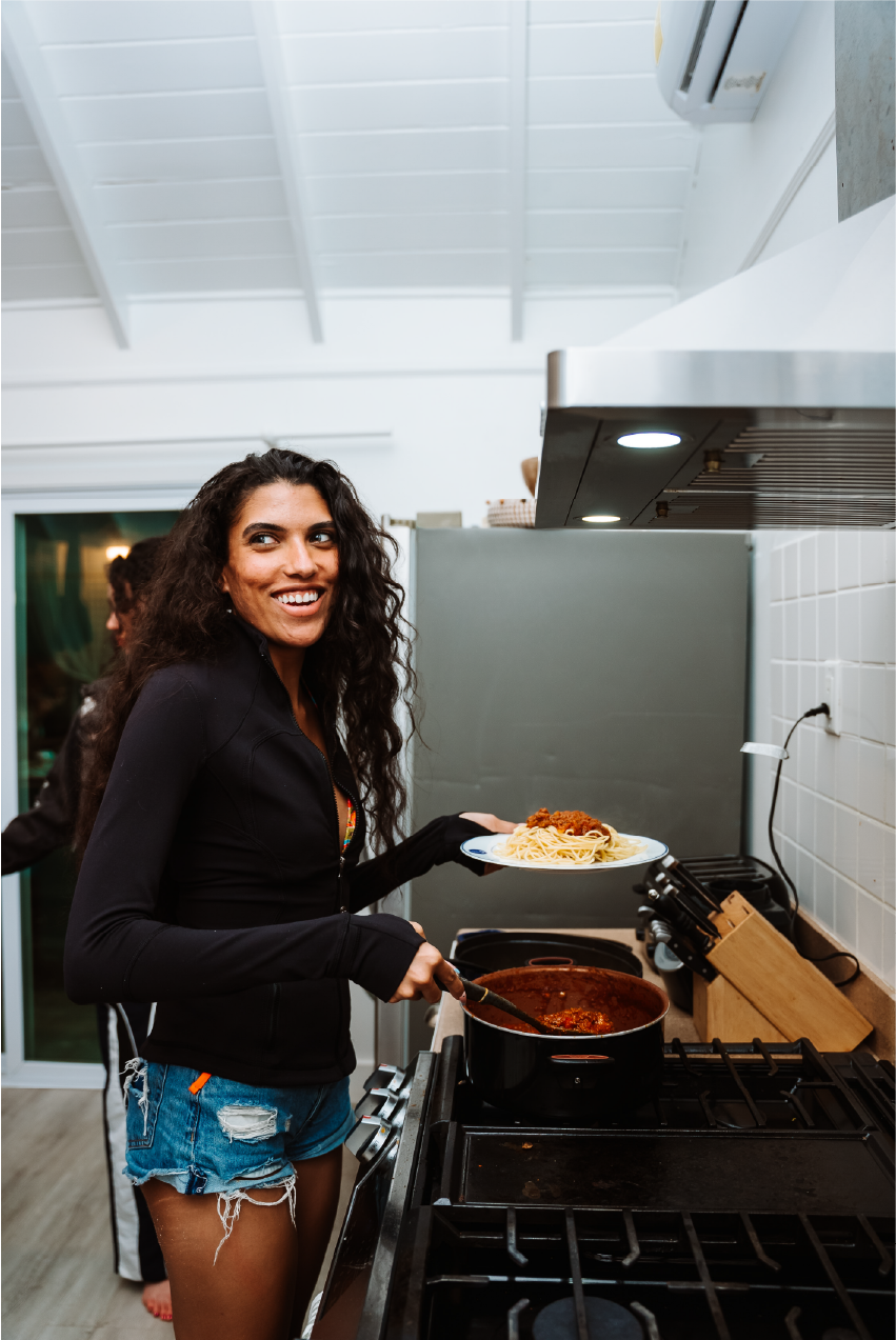 A smiling woman with long curly hair, wearing a black jacket and ripped denim shorts, is cooking pasta with tomato sauce on a stove in a modern kitchen, holding a plate of pasta.