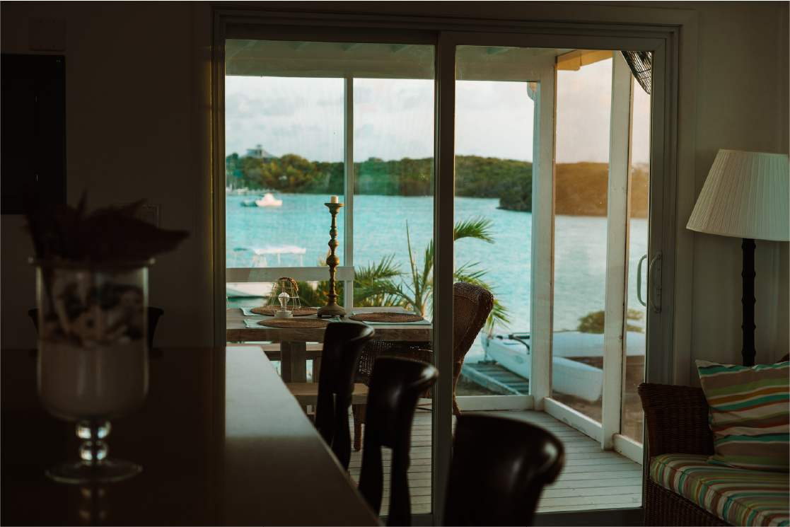 Looking through sliding glass doors at a balcony overlooking water with boats and greenery in the background, inside a cozy living space with a striped armchair, a tall floor lamp, and a dining table with a candle holder.