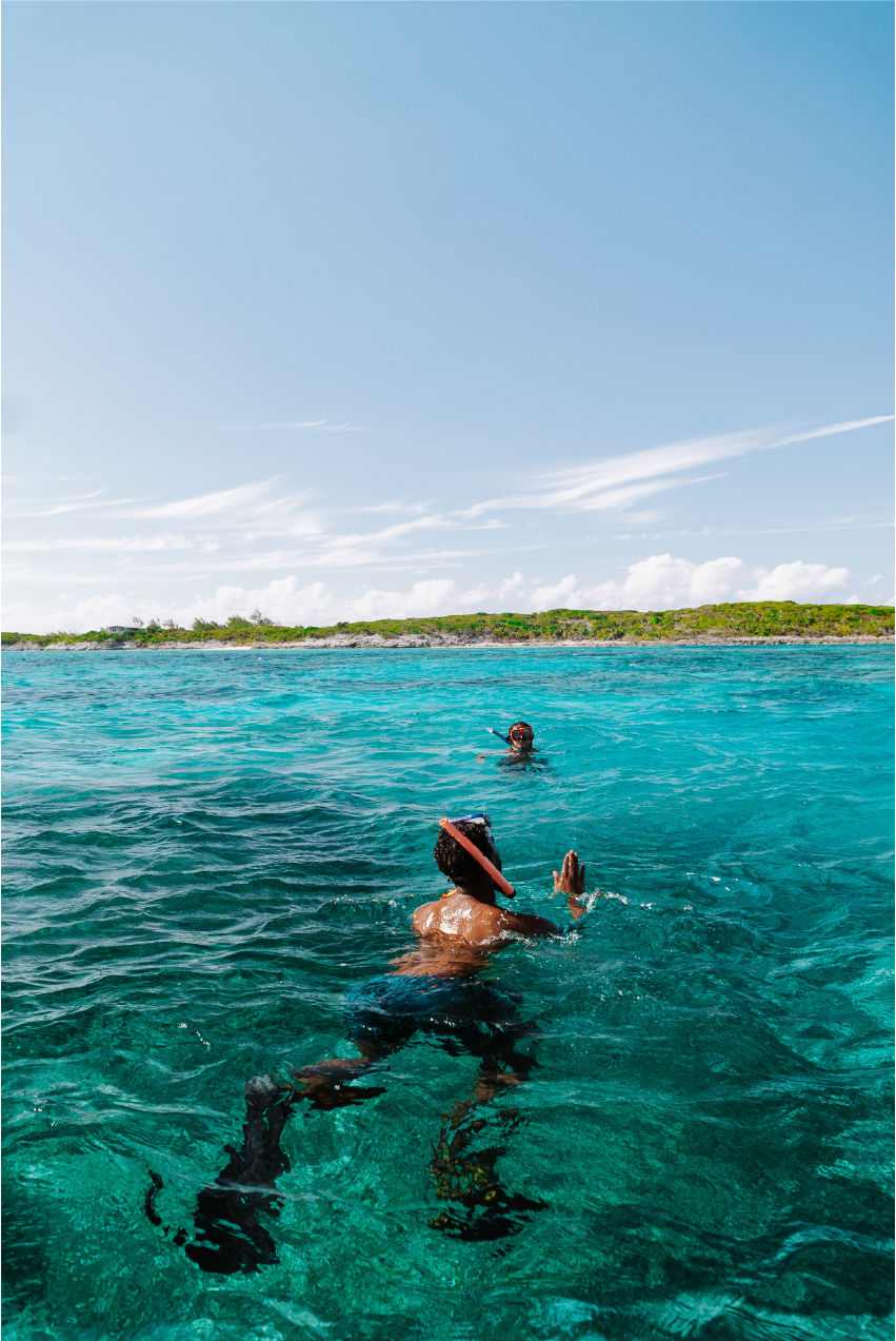 People snorkeling in clear turquoise waters near a coastline with a green landscape under a blue sky with wispy clouds.