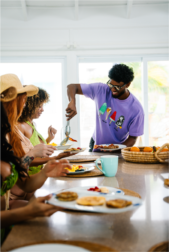 People at a dining table enjoying breakfast with pancakes, syrup, and other foods, with a man serving food and others waiting for their meals.