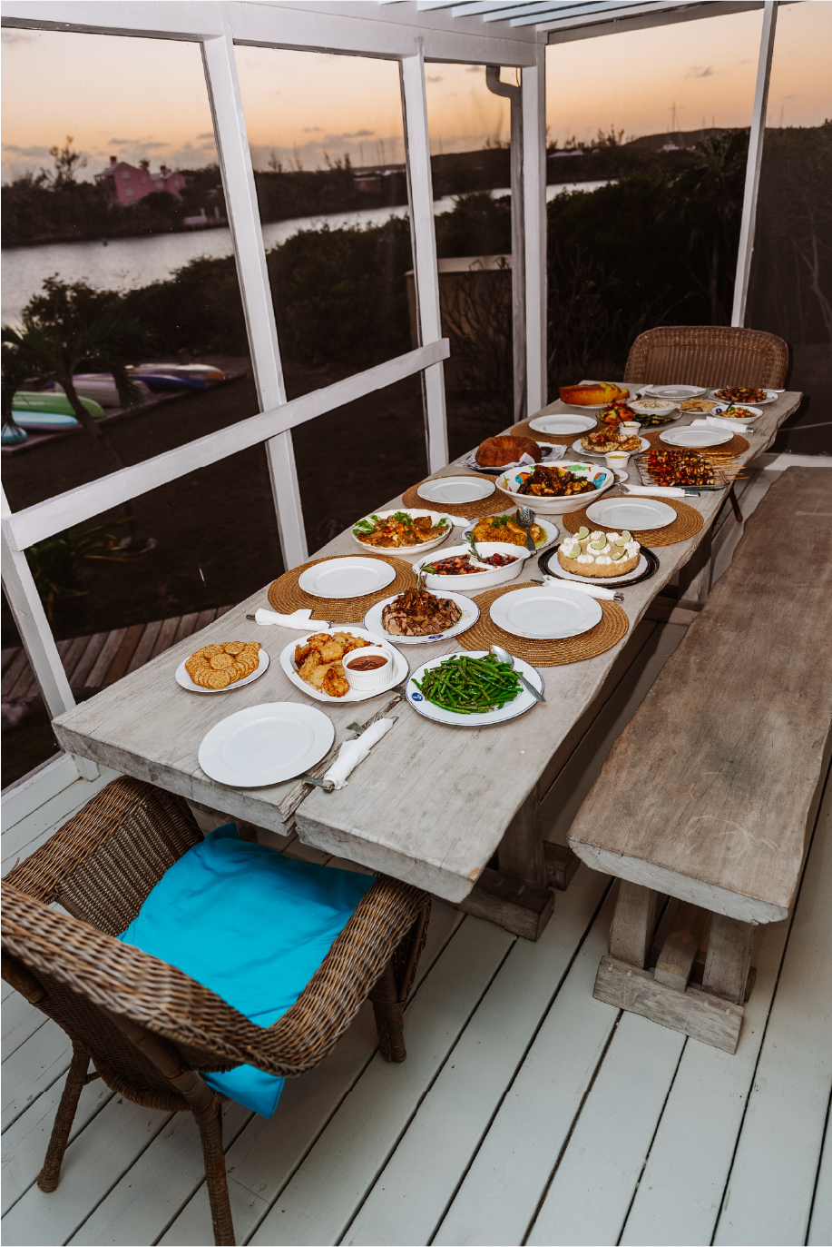 A rustic outdoor dining table set with various dishes, including greens, baked items, desserts, and skewers, on a screened porch overlooking a lake at sunset