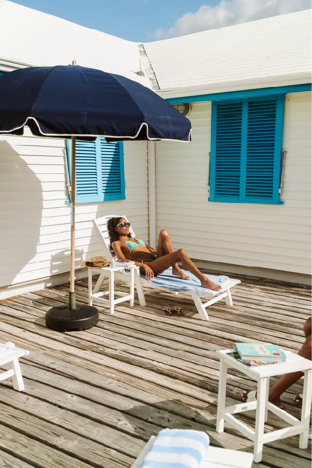 Woman lounging on a deck chair under a large blue umbrella on a wooden patio outside a house with white siding and blue shutters.