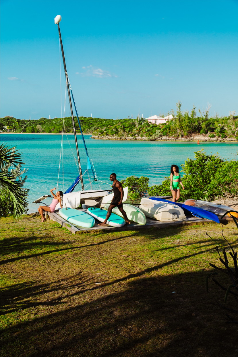 People preparing boats and paddleboards on a dock by a turquoise lake, with two women in bathing suits and a man in casual attire, surrounded by green bushes and trees under a blue sky.