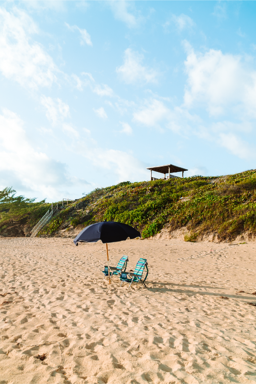 Beach scene with two striped lounge chairs under a black umbrella on sandy shore, green hill with a wooden staircase leading up and a small pavilion on top, blue sky with some clouds.