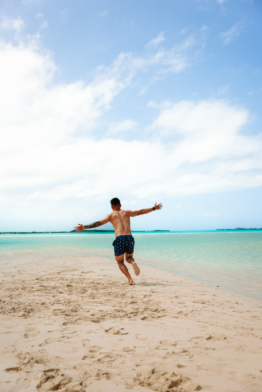 A man in swim trunks running on a sandy beach with open arms, near the shoreline, under a partly cloudy sky.