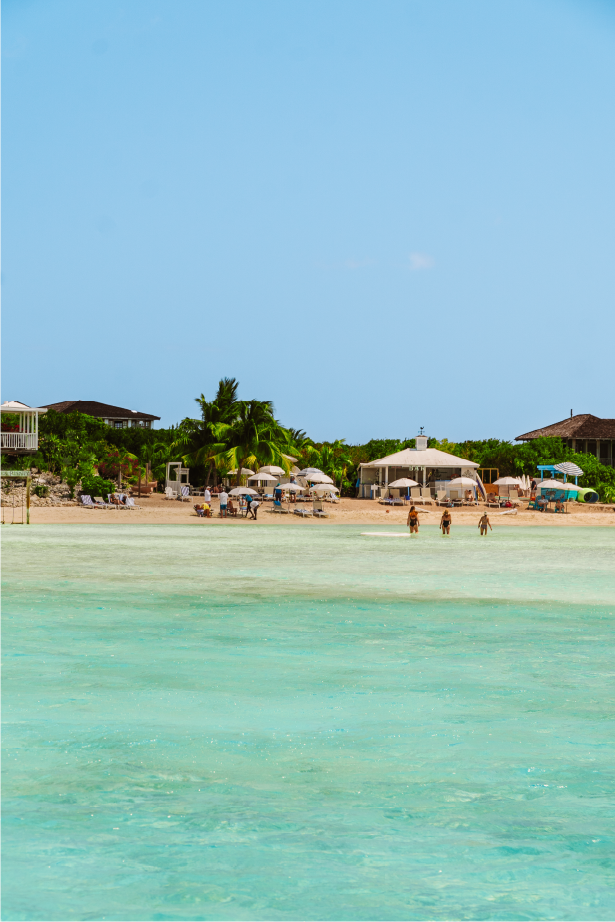 A beach scene with clear turquoise water, sandy shore, palm trees, and beach umbrellas; a few people are walking in the water and on the beach, with houses and umbrellas in the background on a sunny day.
