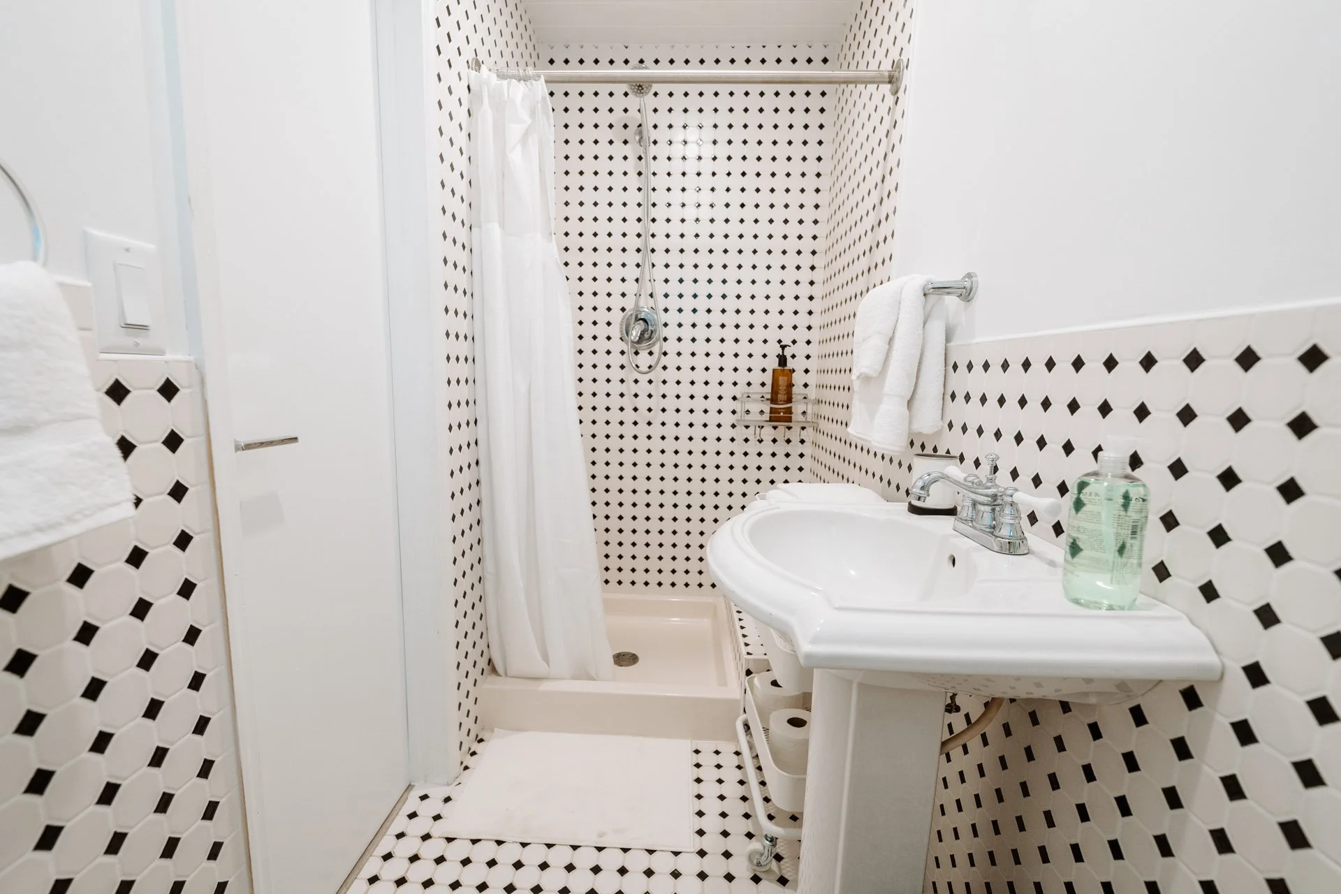 A small bathroom with black and white patterned tiles on the walls and floor, a white shower with a white curtain, a white pedestal sink with a bottle of hand soap, a mirror, and a towel rack with white towels.