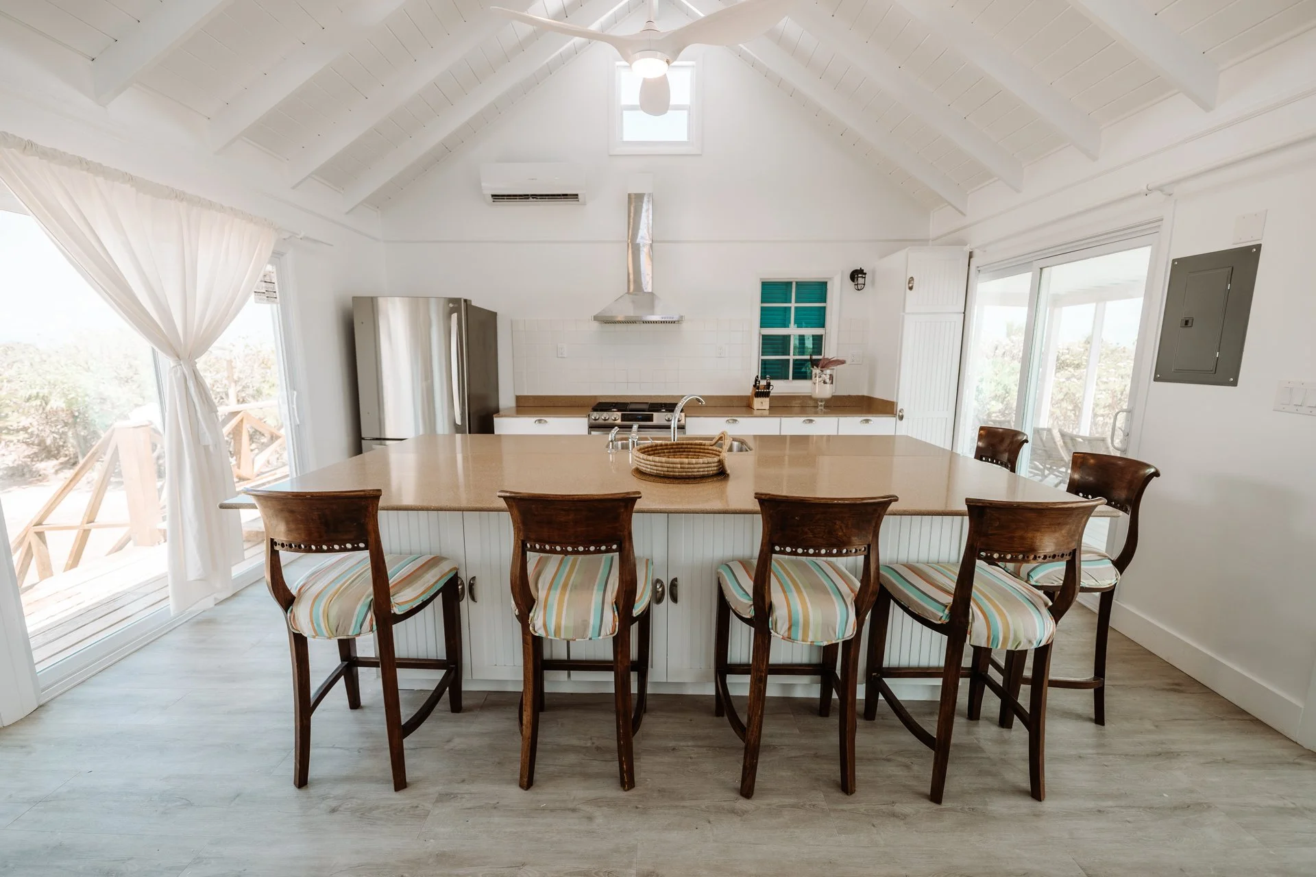 Bright kitchen with high vaulted white ceiling, island with barstools, stainless steel refrigerator, stove with exhaust hood, white tiled backsplash, small window, sliding glass door, and wooden countertops.