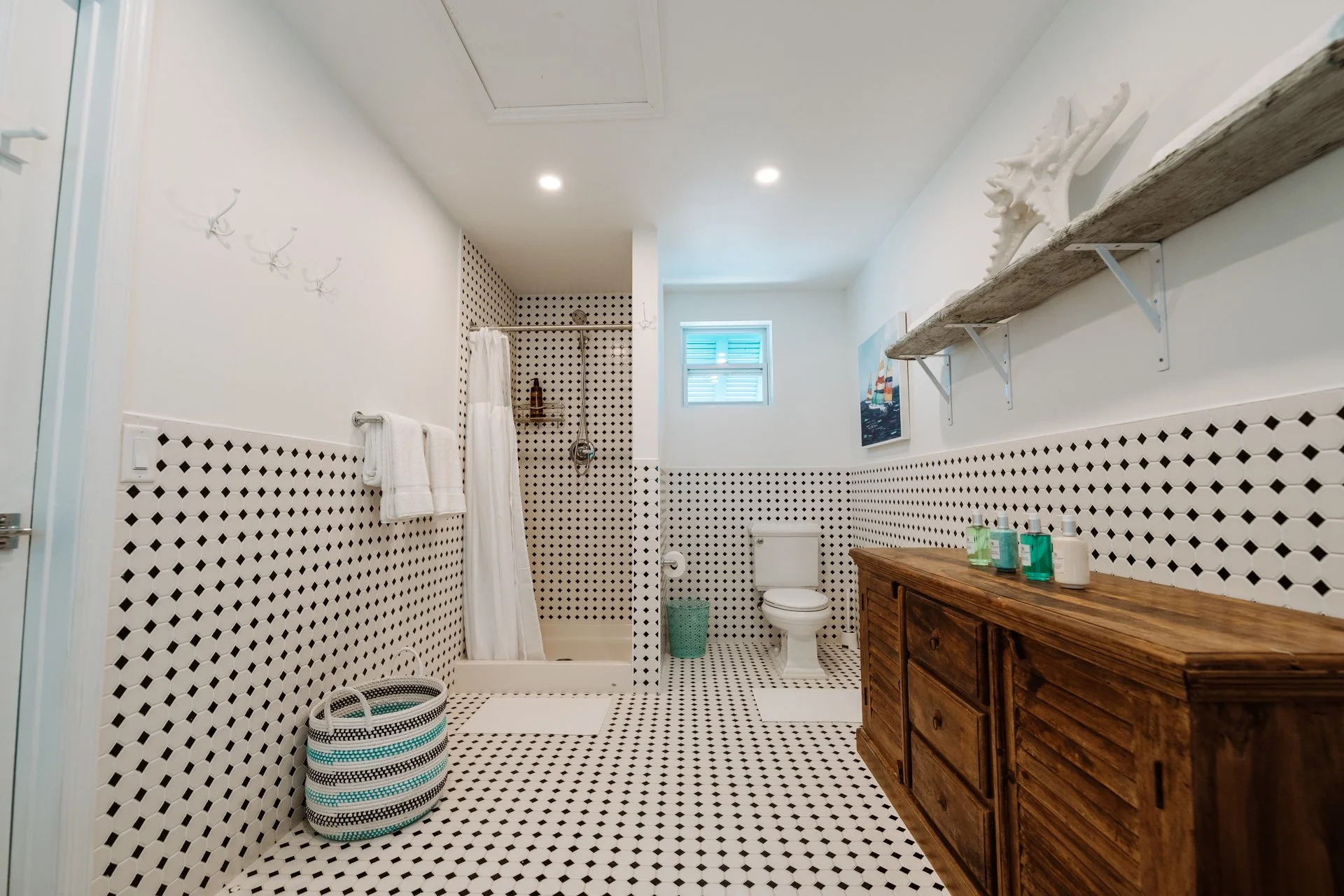 Bathroom with black and white diamond-patterned tiles on the floor and walls, a wooden cabinet, a shower with a white curtain, a toilet, a small window, and a shelf with a decorative white coral piece.
