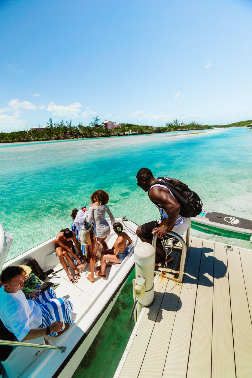 A group of people, including children, on a boat docked at a tropical beach with clear blue water, green island in the background, and a bright blue sky.