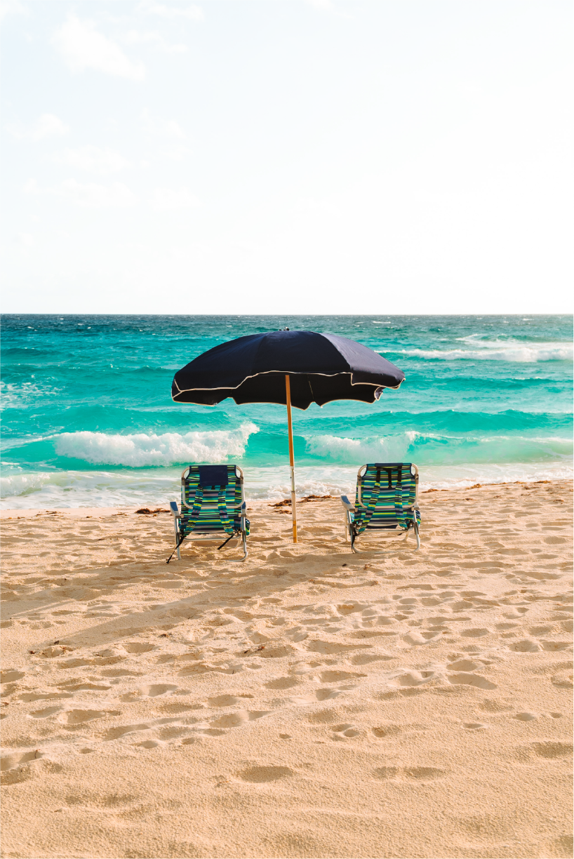 Beach with two lounge chairs and a large black umbrella facing the ocean, with waves and a partly cloudy sky in the background.
