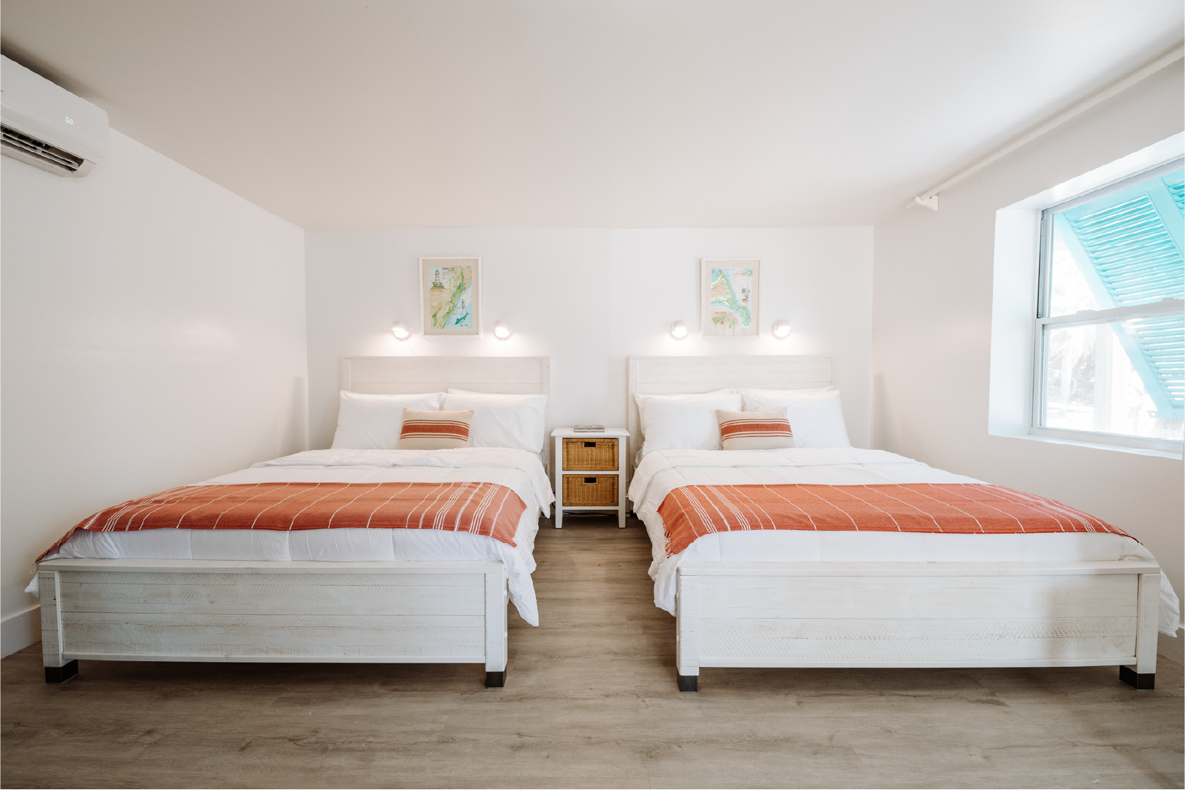 Two white beds with orange striped bed runners and pillows in a bright, minimalist bedroom with wooden flooring, a window with blue shutters, and wall-mounted lights above the headboards.