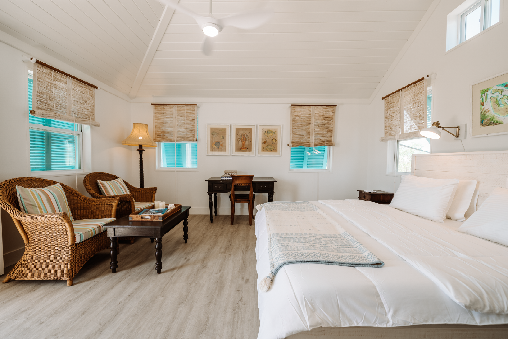Clean and bright bedroom with white walls and ceiling, featuring a large bed with white bedding, a woven armchair and sofa with striped cushions, wooden side tables, a desk with a chair, windows with bamboo shades, and artwork on the walls.