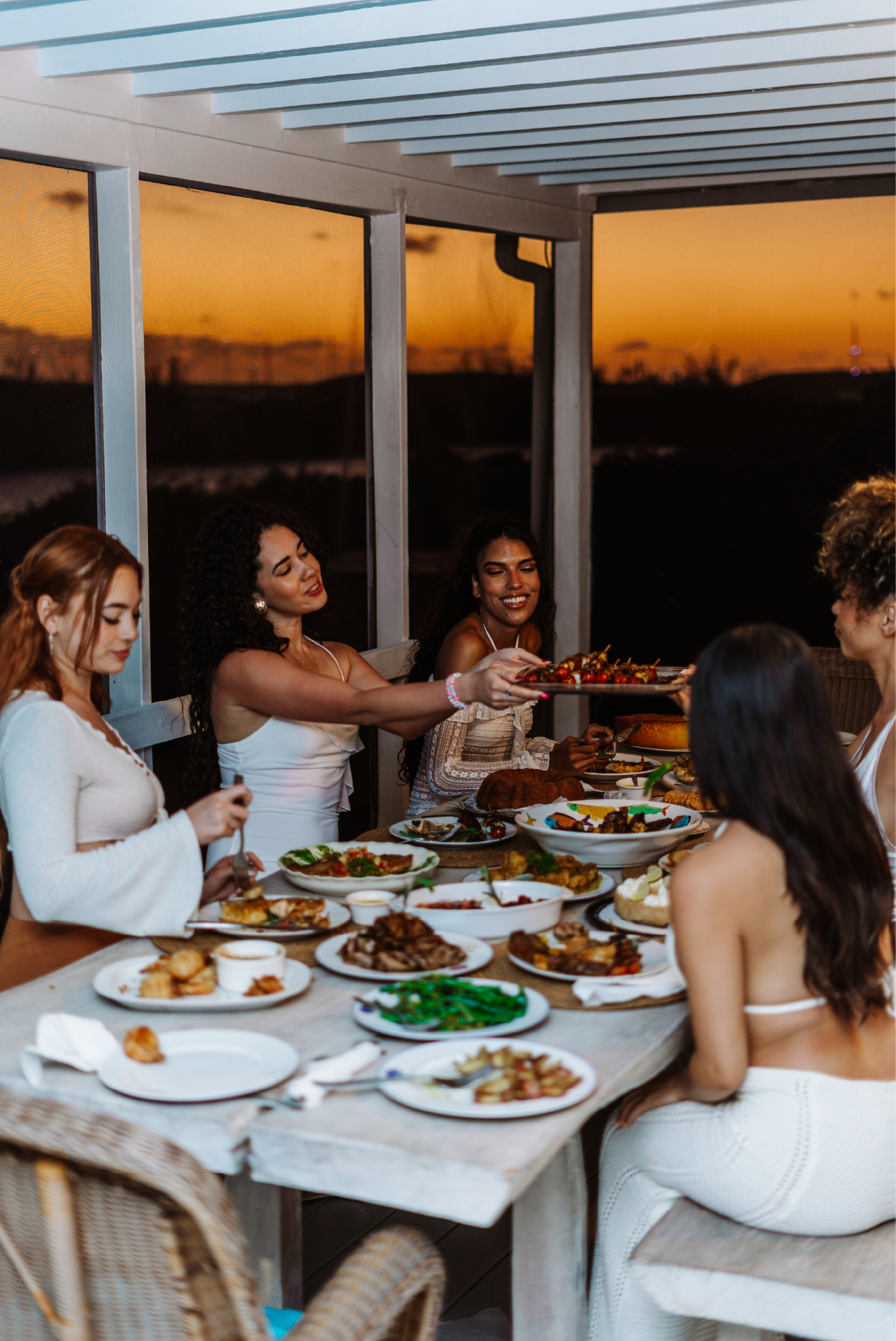 A group of women enjoying dinner at a table filled with various dishes during sunset on a porch.