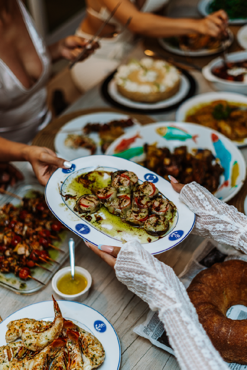 People gather around a table filled with various dishes, with a focus on a plate of grilled shrimp with herbs and oil that one person is handing to another. Other visible foods include skewers, a cake, and a loaf of bread.