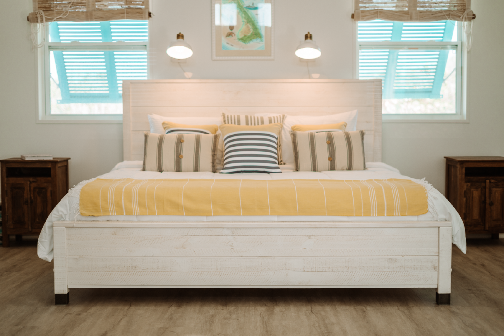 A bedroom with a white wood bed frame, yellow and striped bedding, striped and yellow pillows, two windows with shutters, and two wall-mounted lamps.