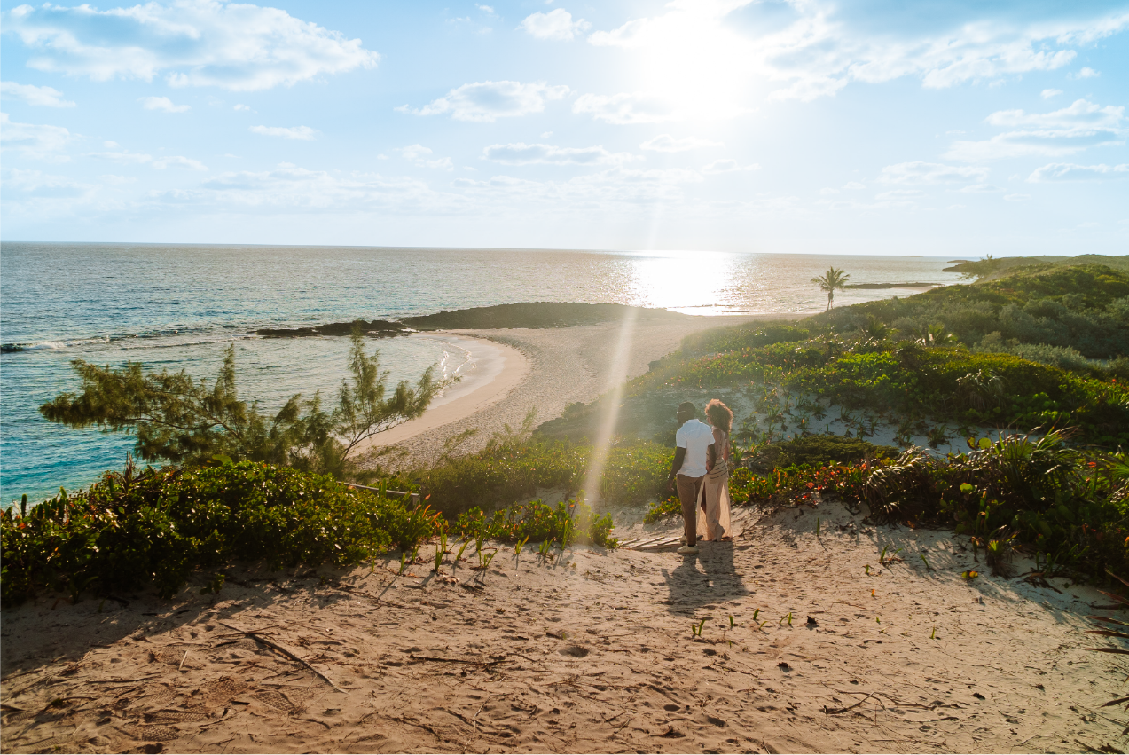 A couple walking down a sandy path towards a beach with clear blue water, some vegetation, and a few palm trees on a sunny day.