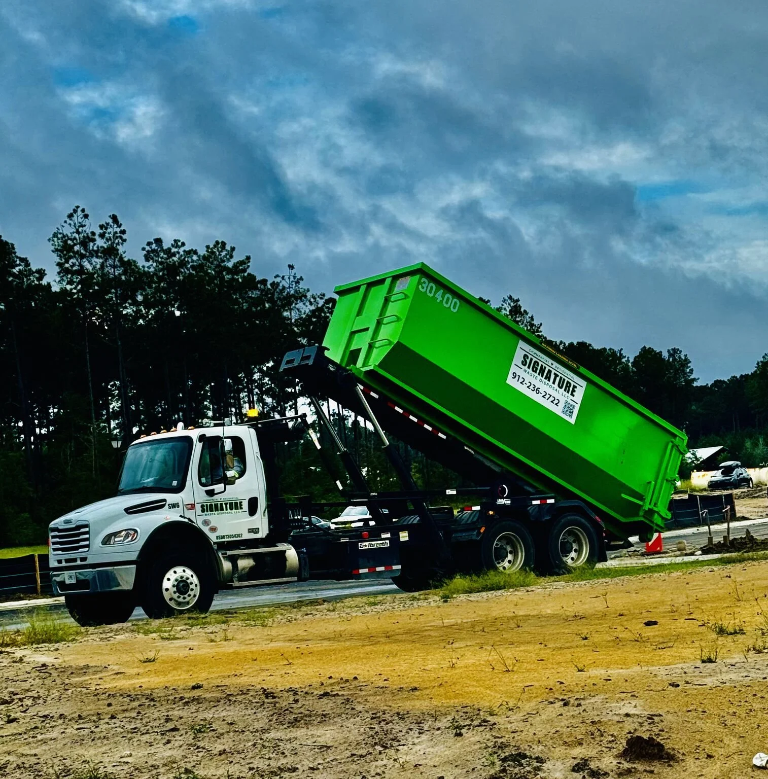A white trash collection truck with a green dumpster raised at a trash transfer station, with trees and a cloudy sky in the background.