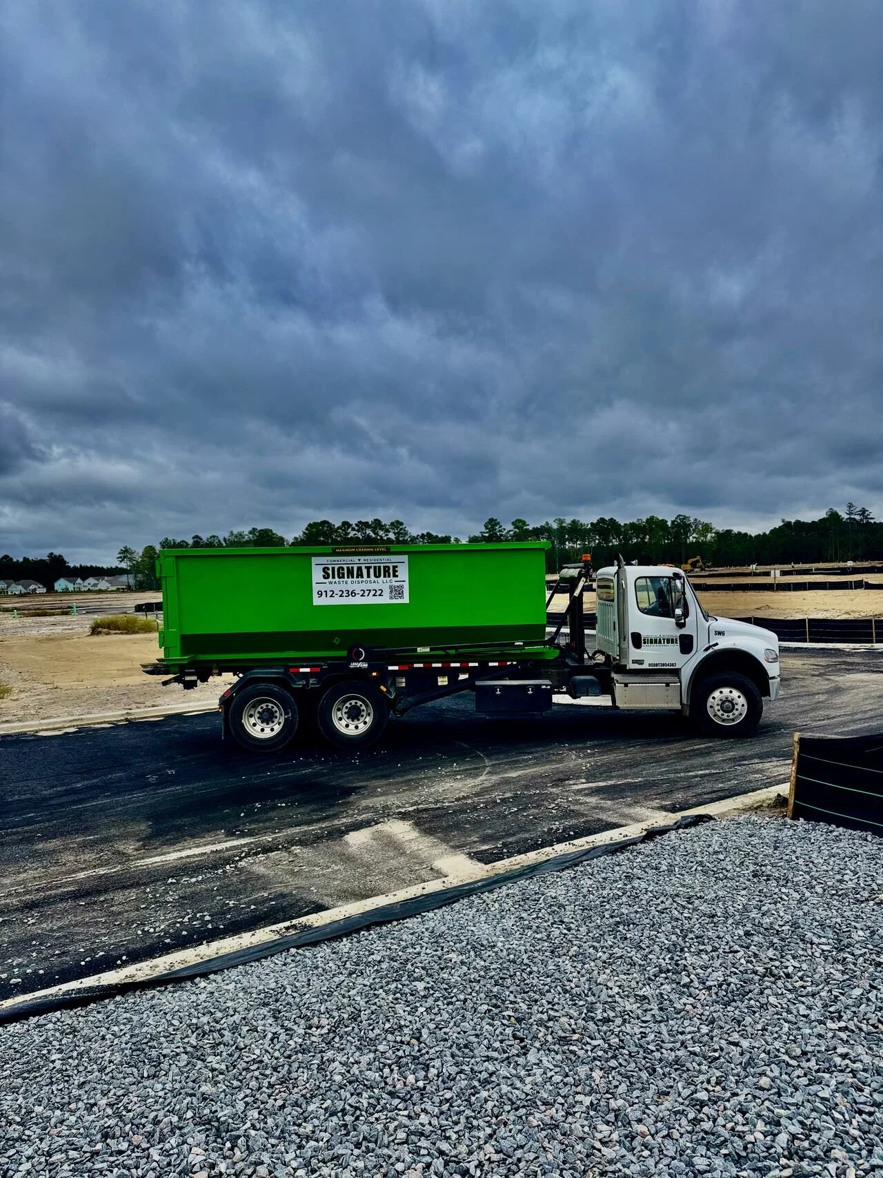 A white dump truck with a green waste disposal bin on its bed parked on a construction site with overcast skies and a dirt landscape.
