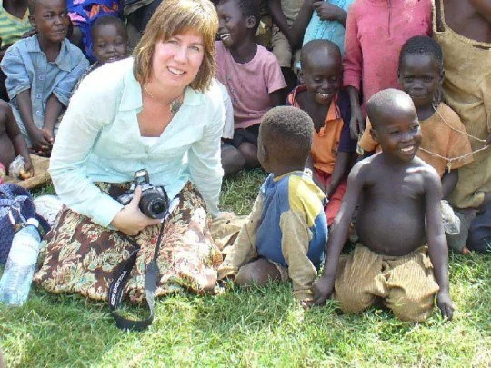 A woman kneeling on grass with a camera, surrounded by smiling children of African descent.