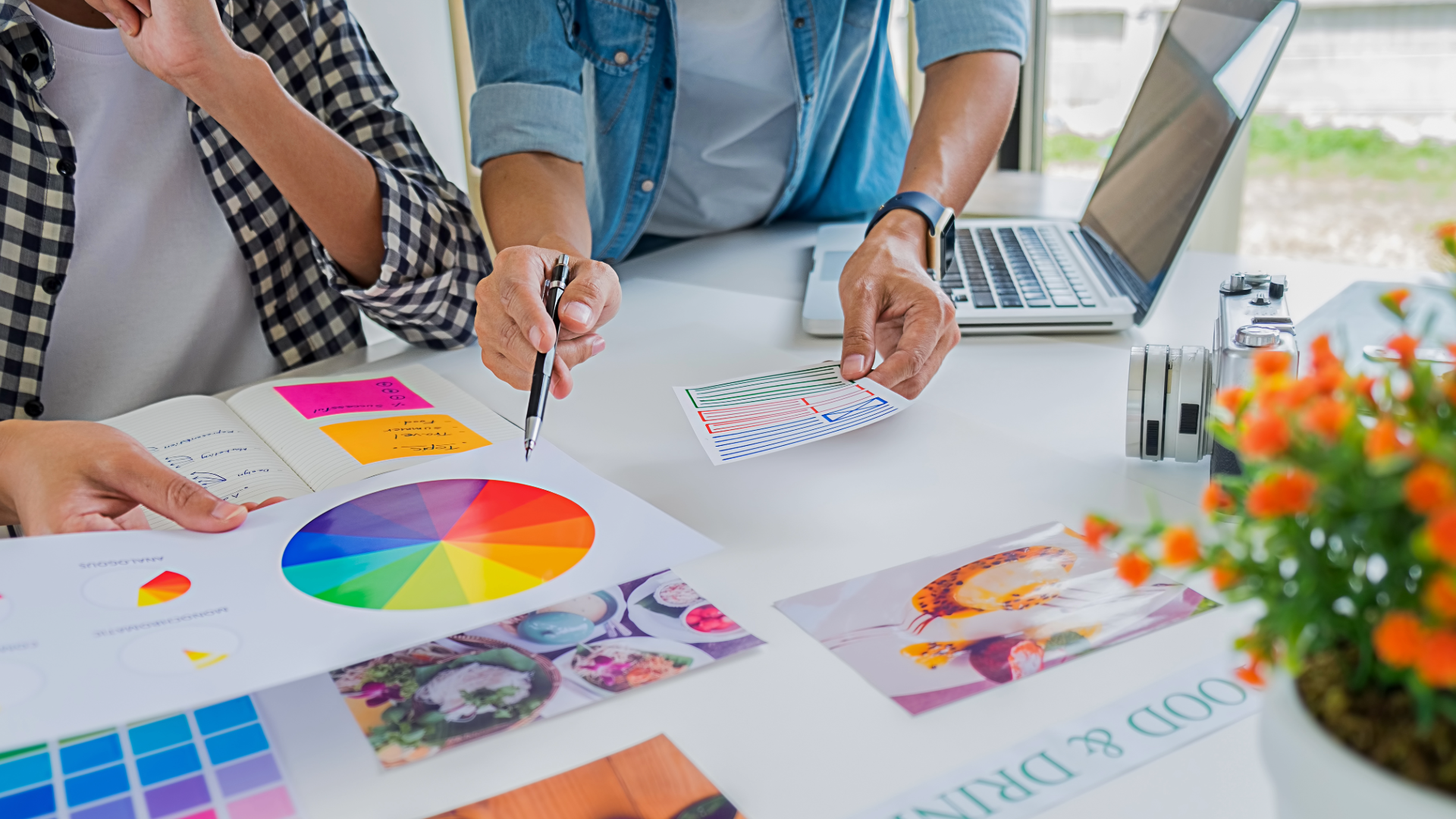 Two people working at a table with color charts, notes, and a laptop, discussing design or branding, with a potted plant in the foreground.