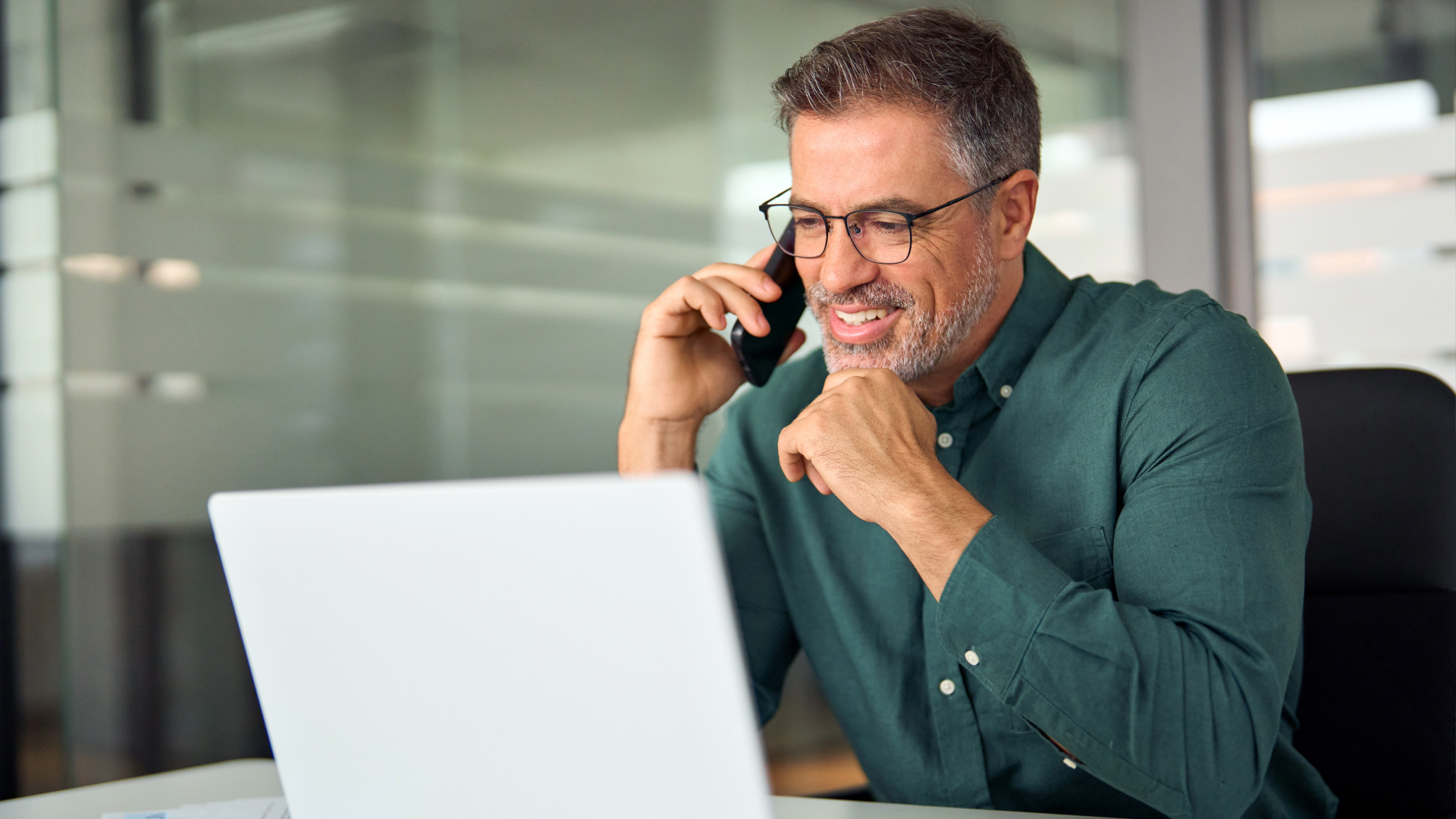 Middle-aged man with glasses and a beard, talking on a mobile phone while working on a laptop in an office.