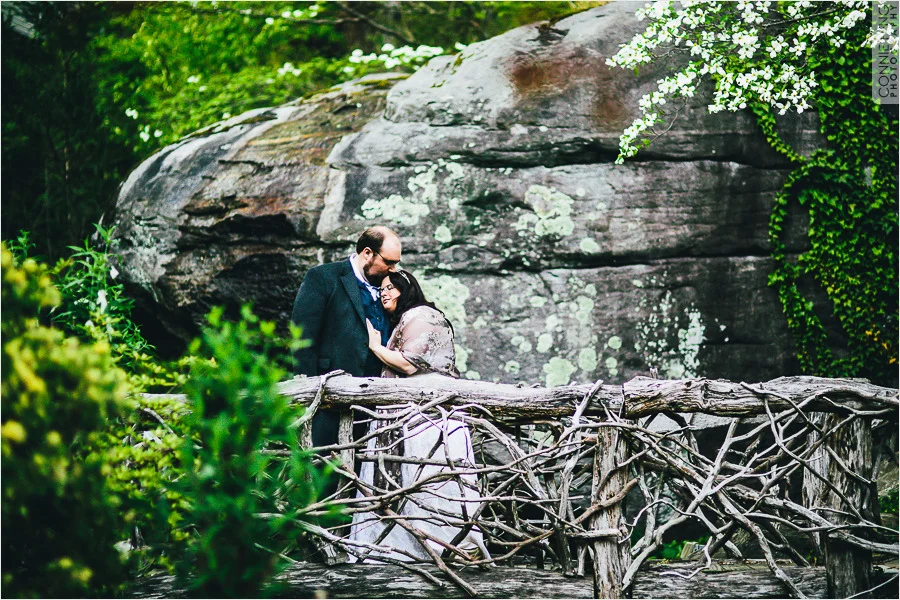 A couple embraces on a rustic wooden bridge in a lush green forest, with a large rock and white flowers in the background.