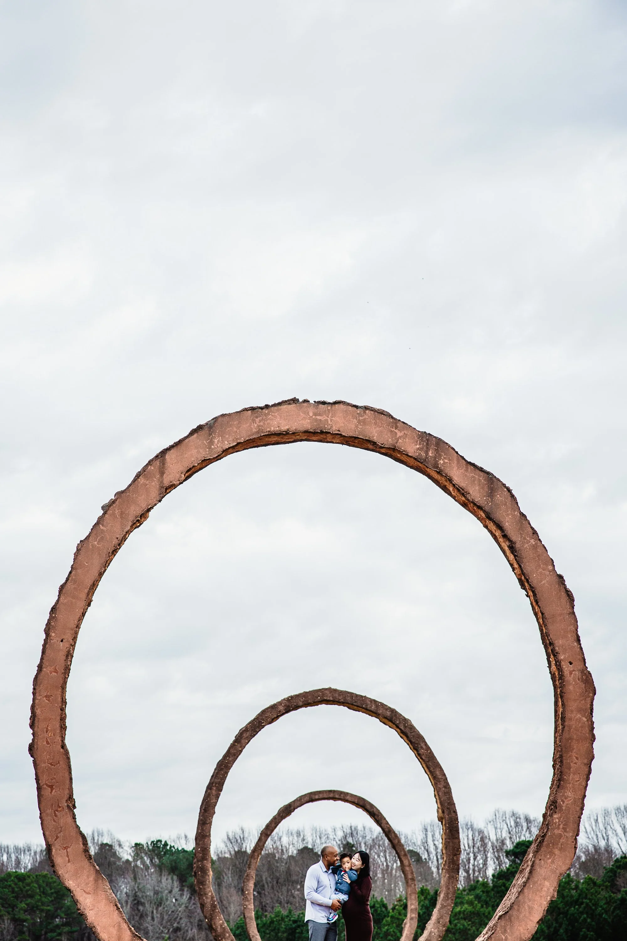 A family of three standing outdoors among large circular metal structures, with overcast sky and trees in the background at NC Museum of Art.