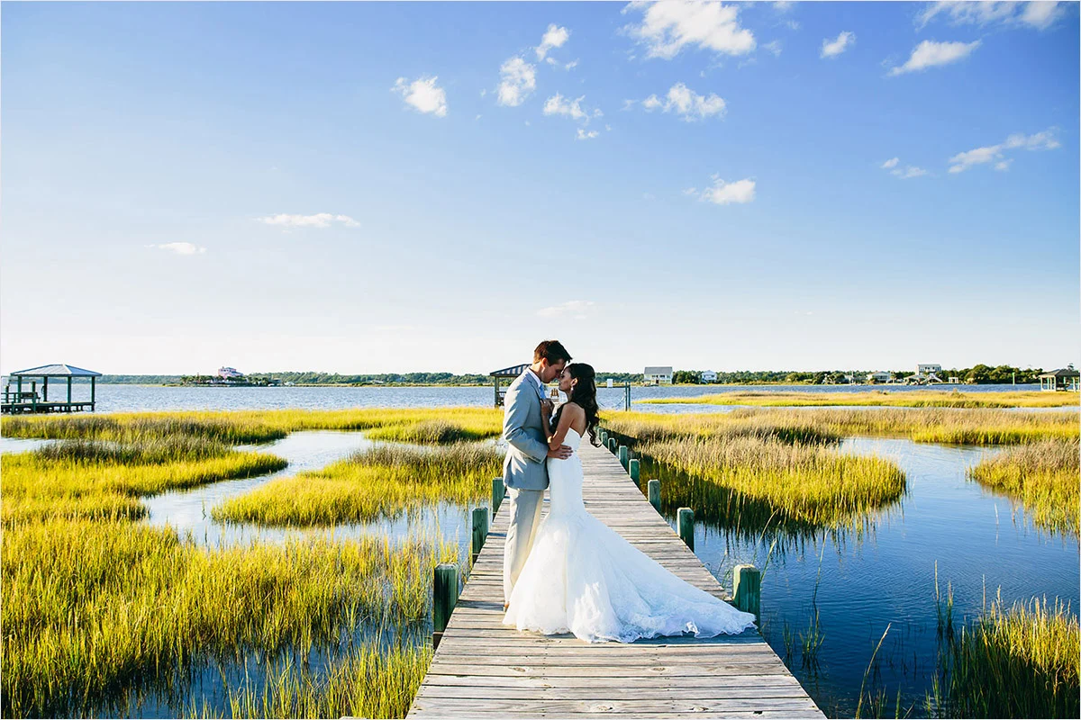 A bride and groom embrace on a wooden dock over marshland during a sunny day, with blue skies and water surrounding the area.