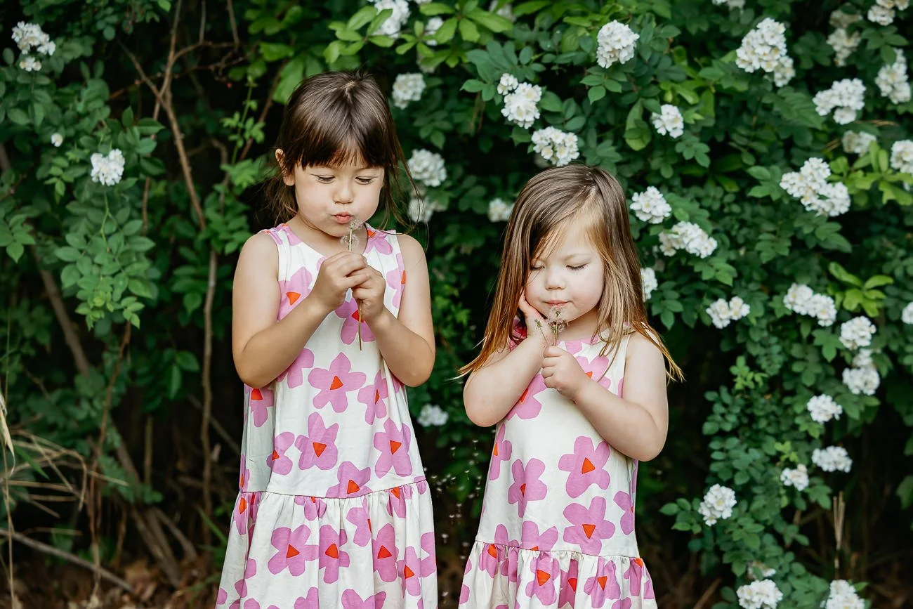 Two young girls in matching pink and white floral dresses smelling flowers in a garden with white blooms and green foliage.