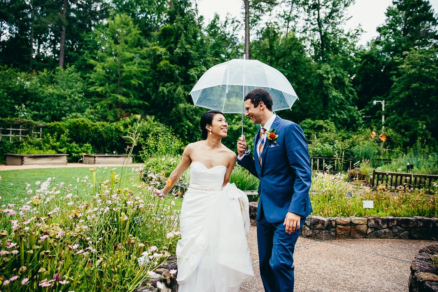 A bride and groom on their wedding day, sharing a joyful moment under a transparent umbrella in a lush garden, with the bride smiling at the groom and the bride's wedding dress and groom's blue suit visible.