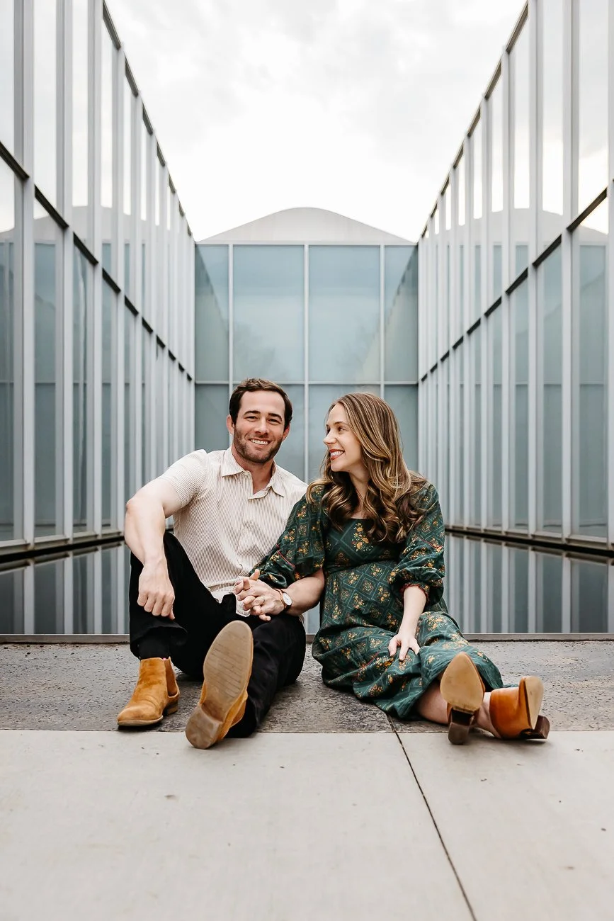 A smiling man and woman sitting on the ground holding hands in a modern, glass-walled courtyard.