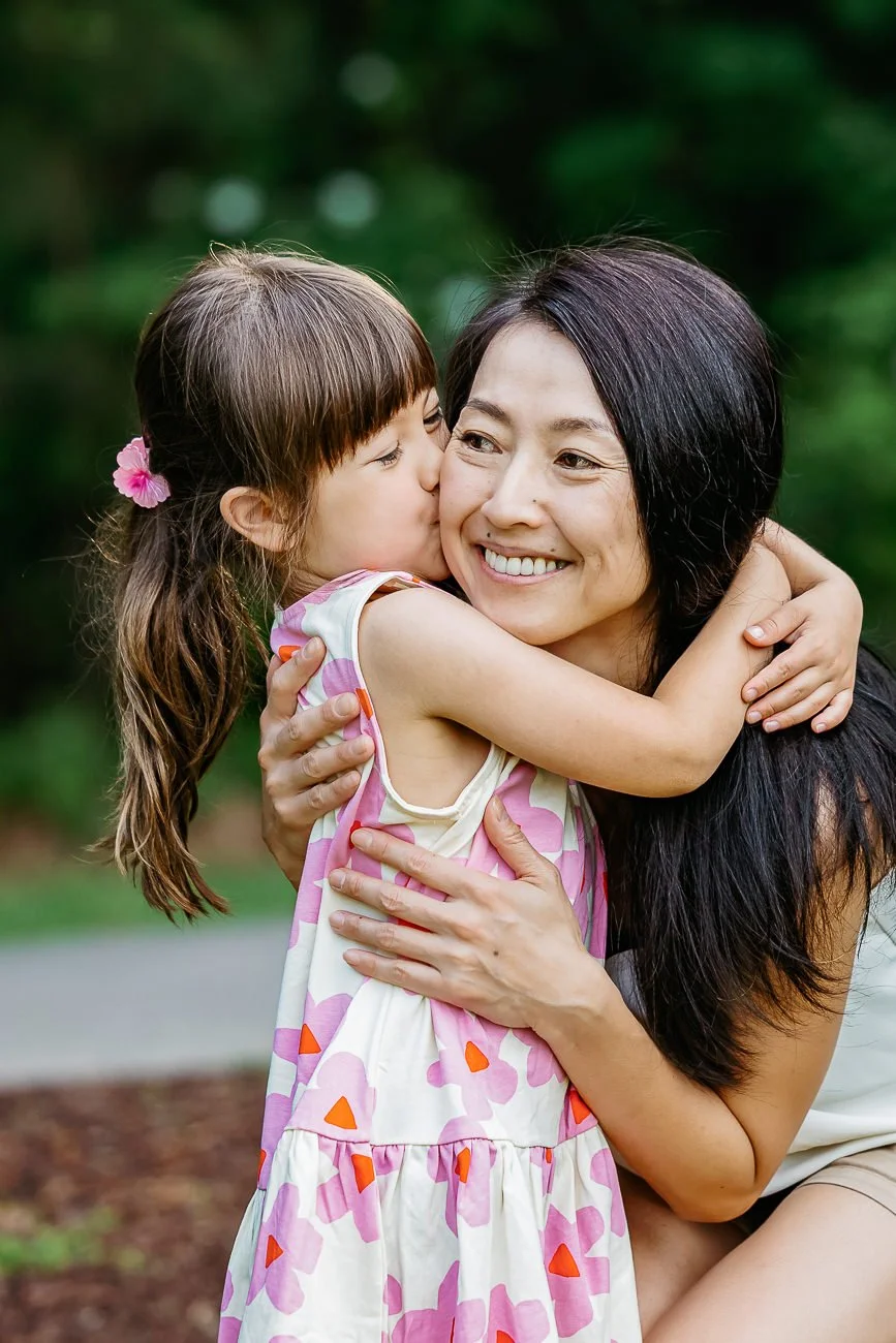A young girl with brown hair and pink flowers in her hair hugging and kissing an Asian woman with black hair, both smiling warmly outdoors with green trees in the background.