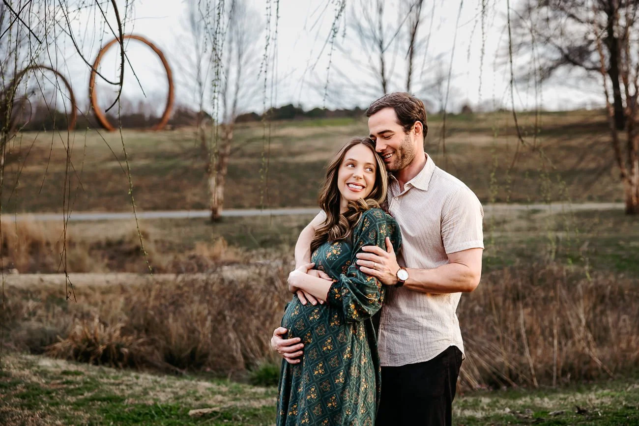 A couple hugging outdoors in a park with trees and a large wooden heart sculpture in the background.