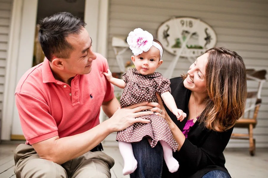A smiling man and woman holding a baby girl outside on a porch.