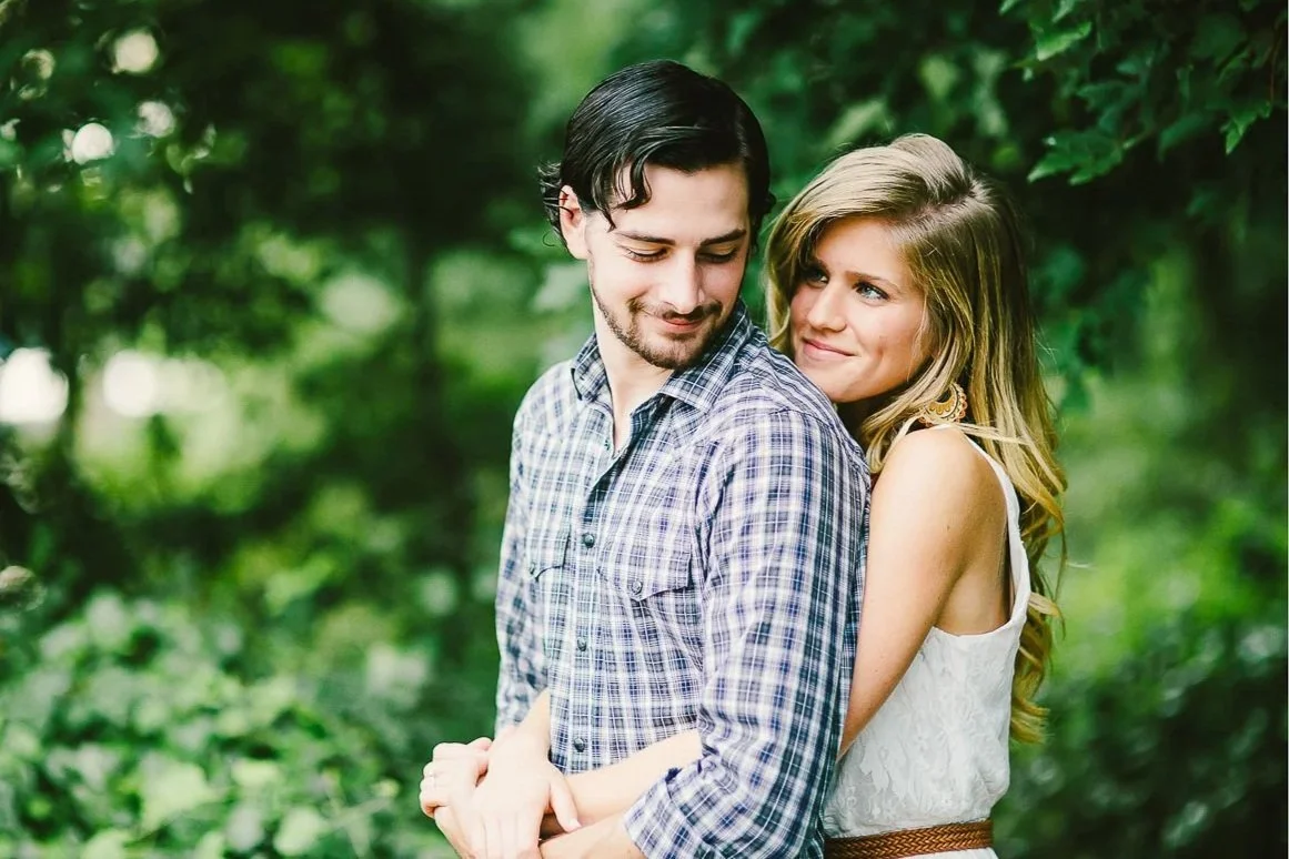 A young couple enjoying a moment outdoors in a lush green forest. The man has dark hair and a beard, wearing a checkered shirt. The woman has long blonde hair, wearing a white sleeveless top and earrings. They are standing close, with the woman embracing the man's back, both with gentle expressions.