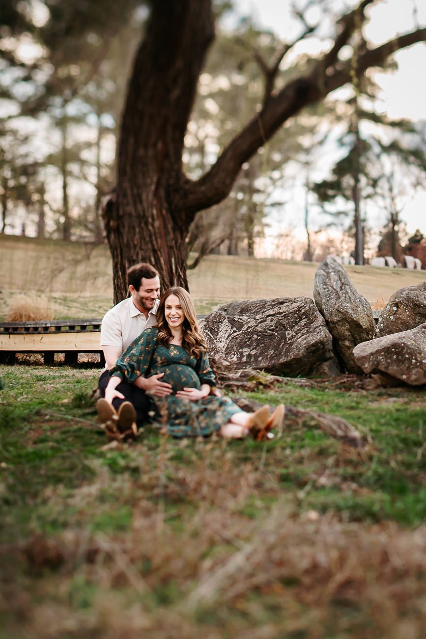 A pregnant woman and a man sitting on the grass in a park, smiling, with the woman resting her hands on her belly, surrounded by rocks and trees.