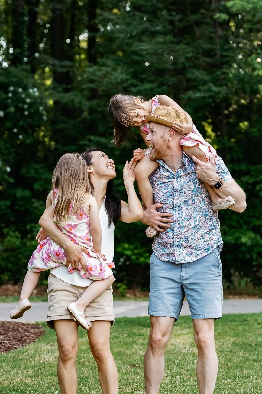 A family of four enjoying outdoor time, with the father carrying a young girl on his shoulders, and the mother holding the other girl, all smiling and engaging with each other in a green park.