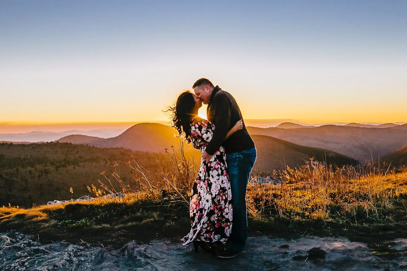 A couple is standing outdoors at sunset, embracing and about to kiss, with a scenic view of mountains and a colorful sky in the background.