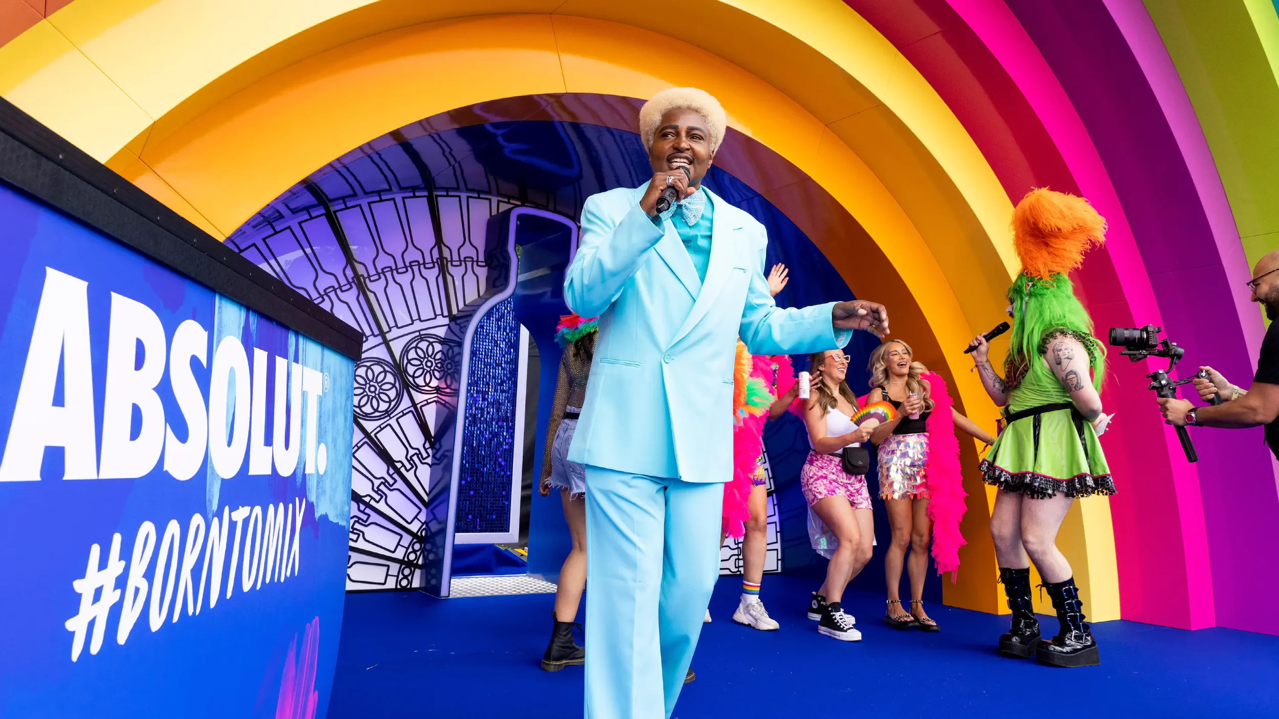A group of people on a colorful stage with rainbow arches. There is a man in a light blue suit holding a microphone, and a woman with bright orange and green hair wearing a green dress and black boots. Several people are laughing and smiling, with one holding a rainbow-colored fan. To the left, there is a large blue sign with the word 'ABSOLUT' and '#BORNONTOP' written on it.