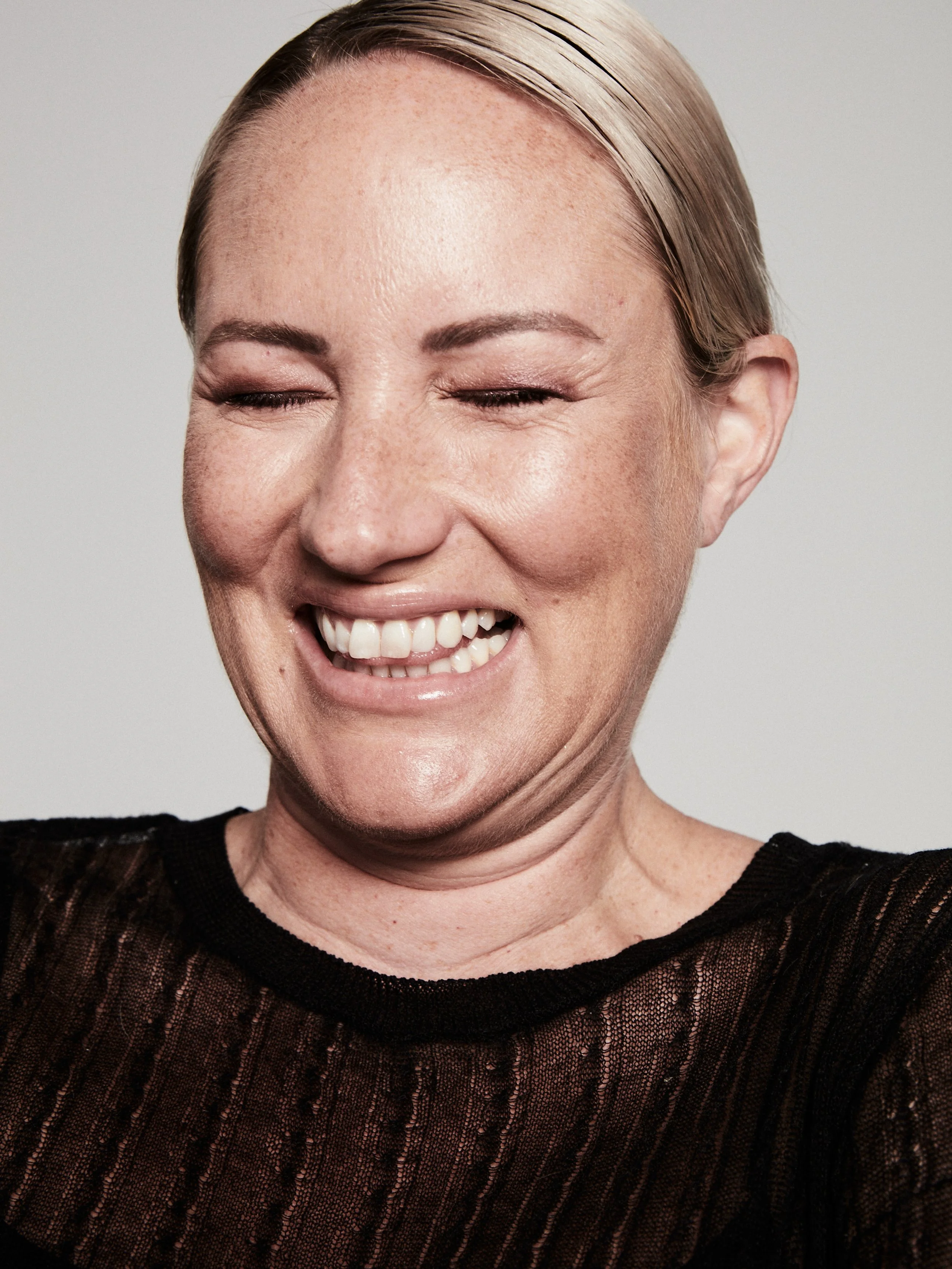 A woman with blonde hair smiling with eyes closed, wearing a black lace top.