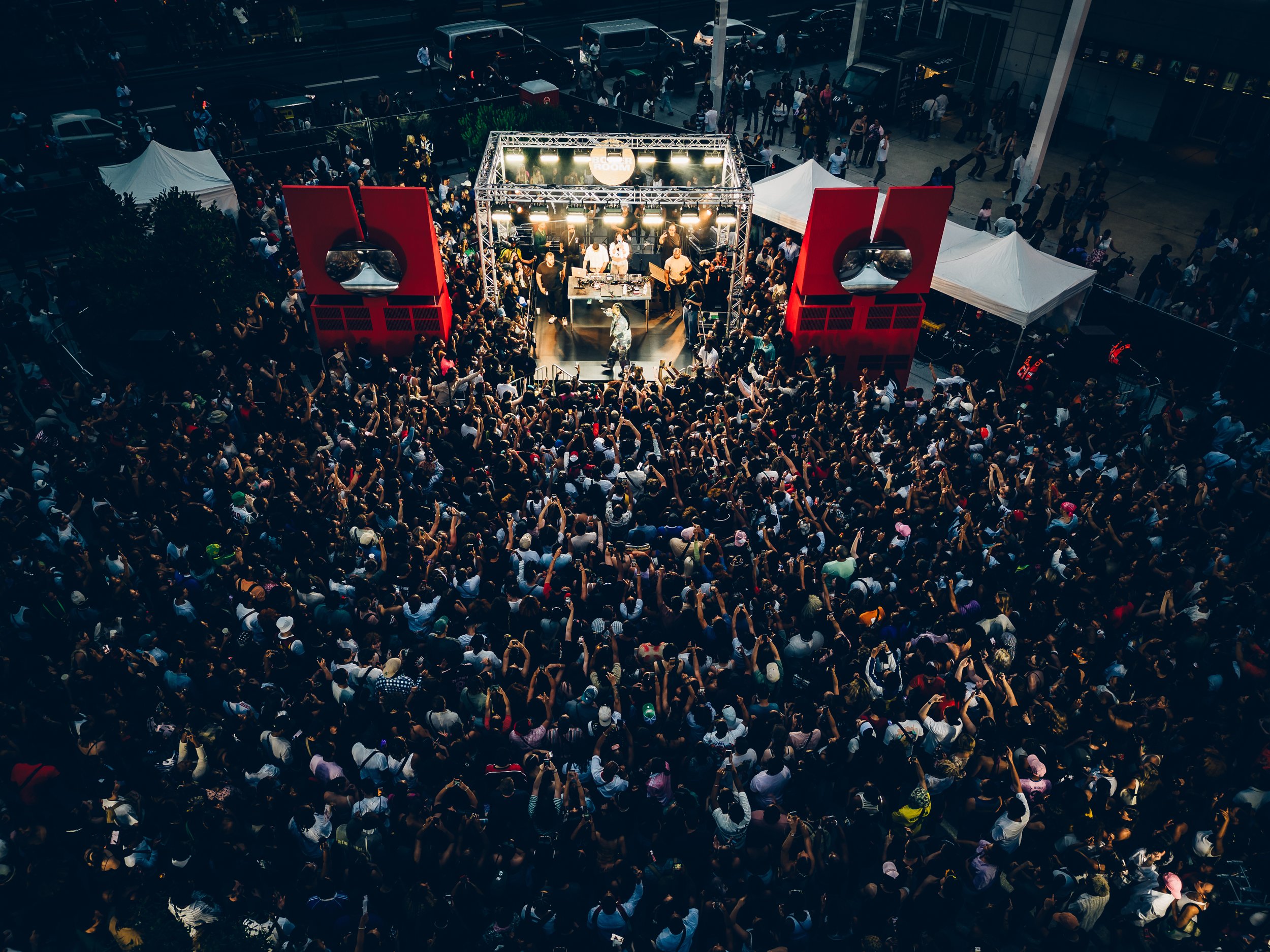Bird's-eye view of a crowded outdoor concert with a band performing on stage, surrounded by large red speakers and lively audience.