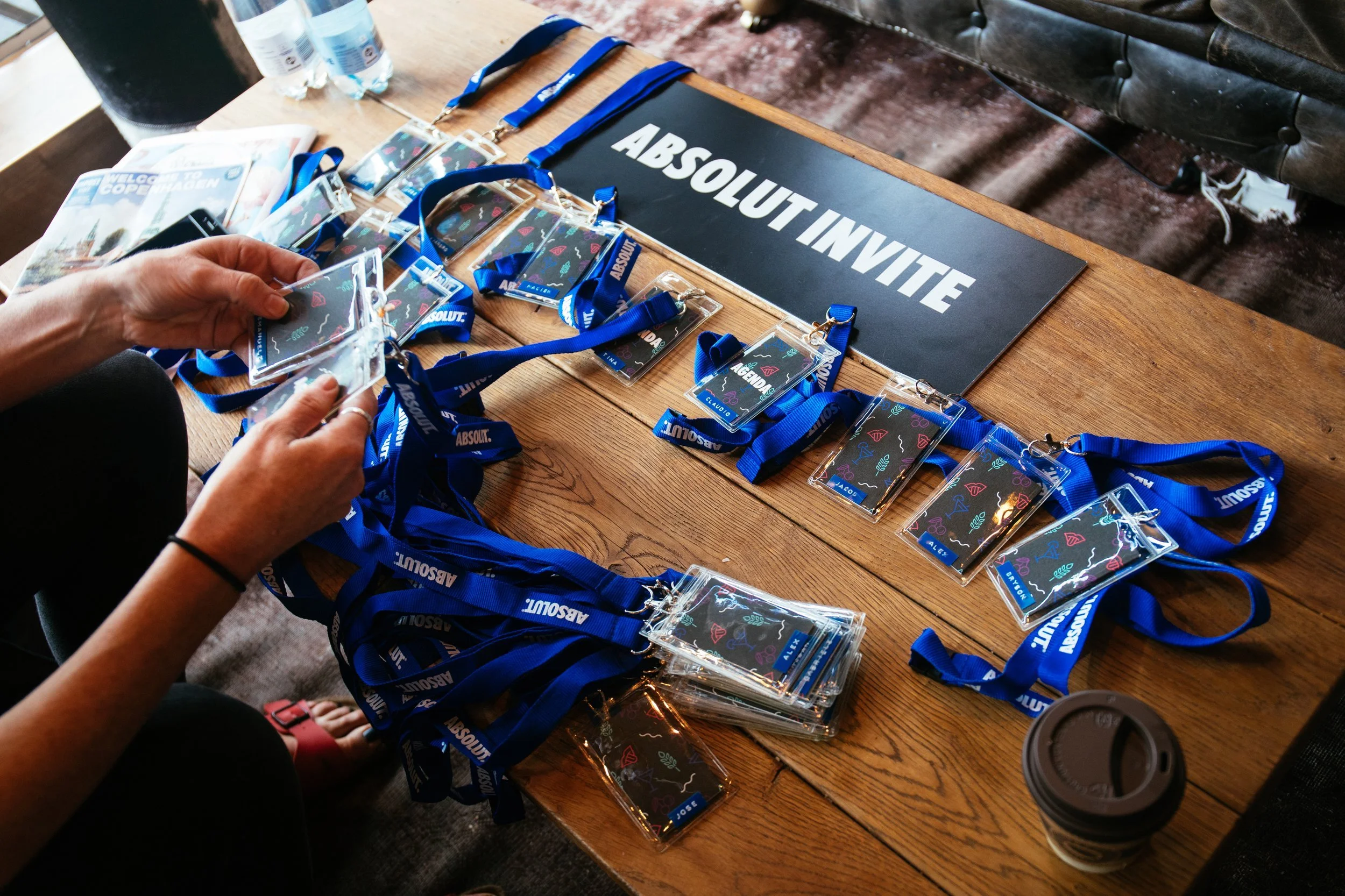 A person working at a wooden table with numerous lanyards and name badges for an event called ABSOLUT INVITE, with a sign displaying the same text, and bottles of water and a coffee cup also on the table.
