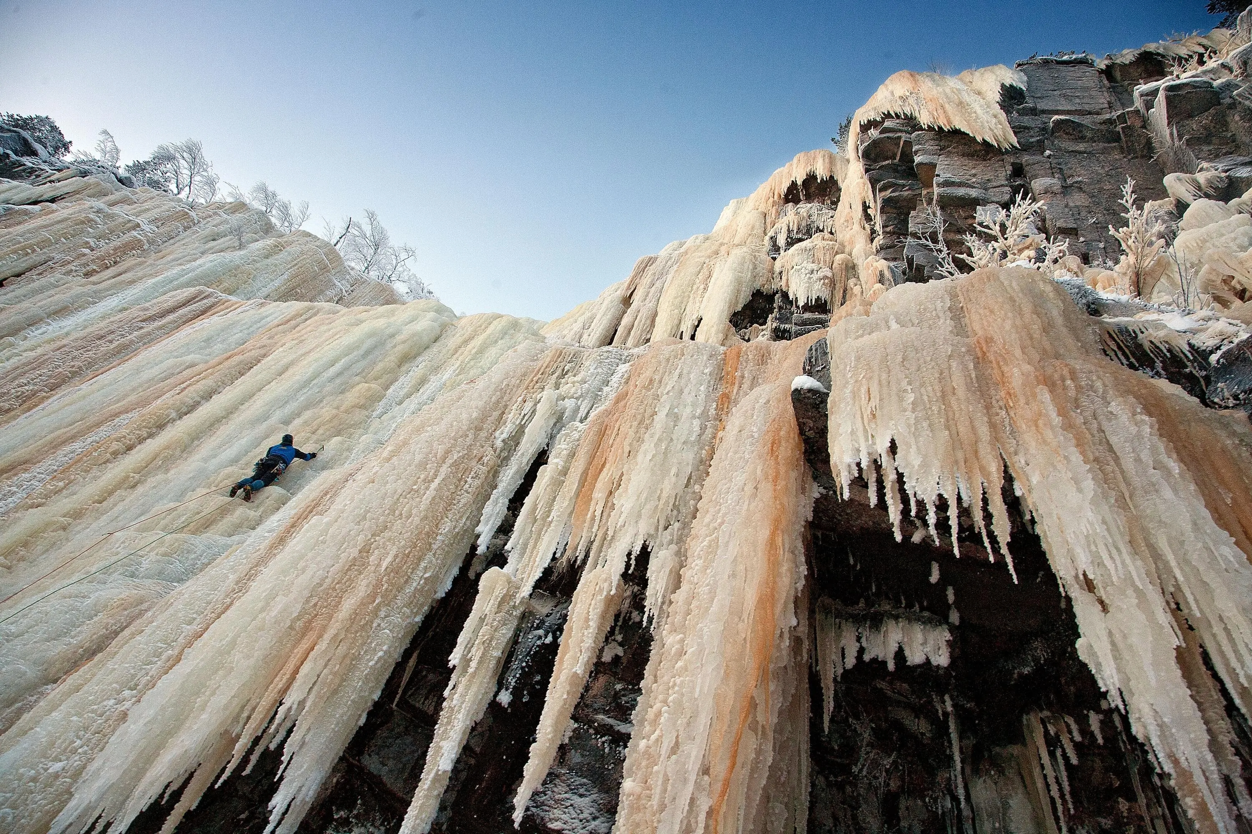 An ice climber ascending a frozen waterfall on a steep, icy cliff with ice formations and snow-covered trees at the top.