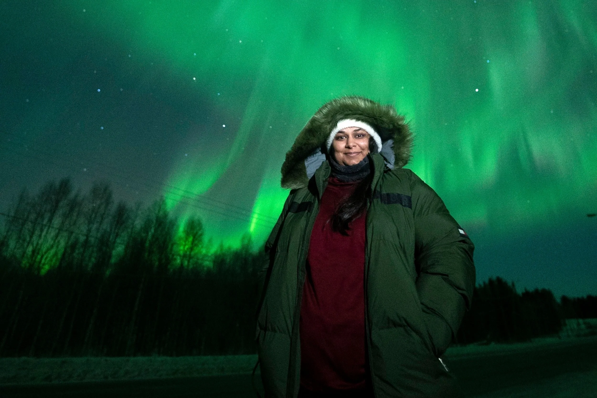 Woman in a green winter coat with a fur-lined hood standing outdoors at night under the Northern Lights with trees in the background.