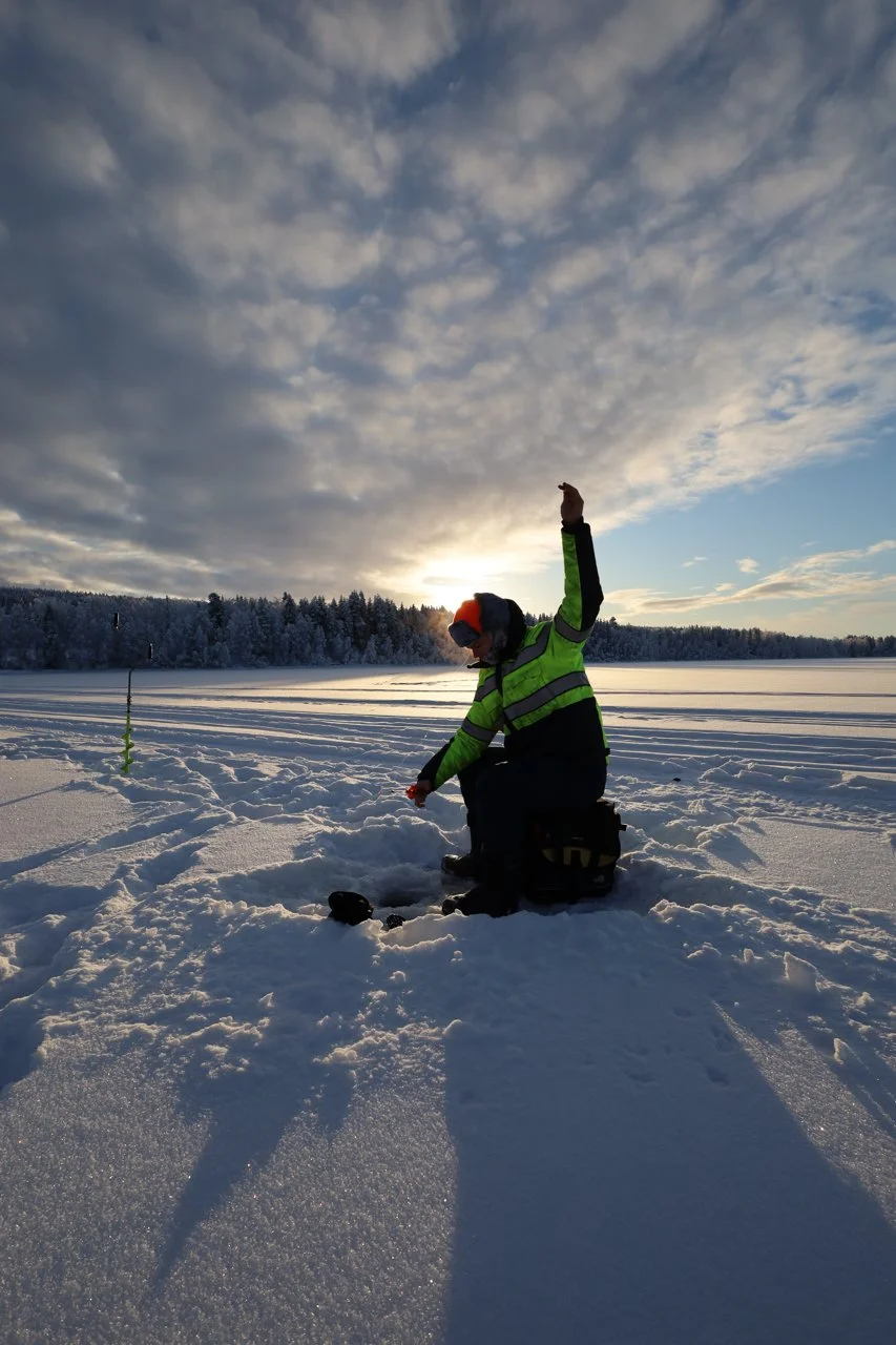 Person dressed in winter gear kneeling in snow on a frozen lake or field, with a forest in the background and a cloudy sky at sunset, raising one arm.