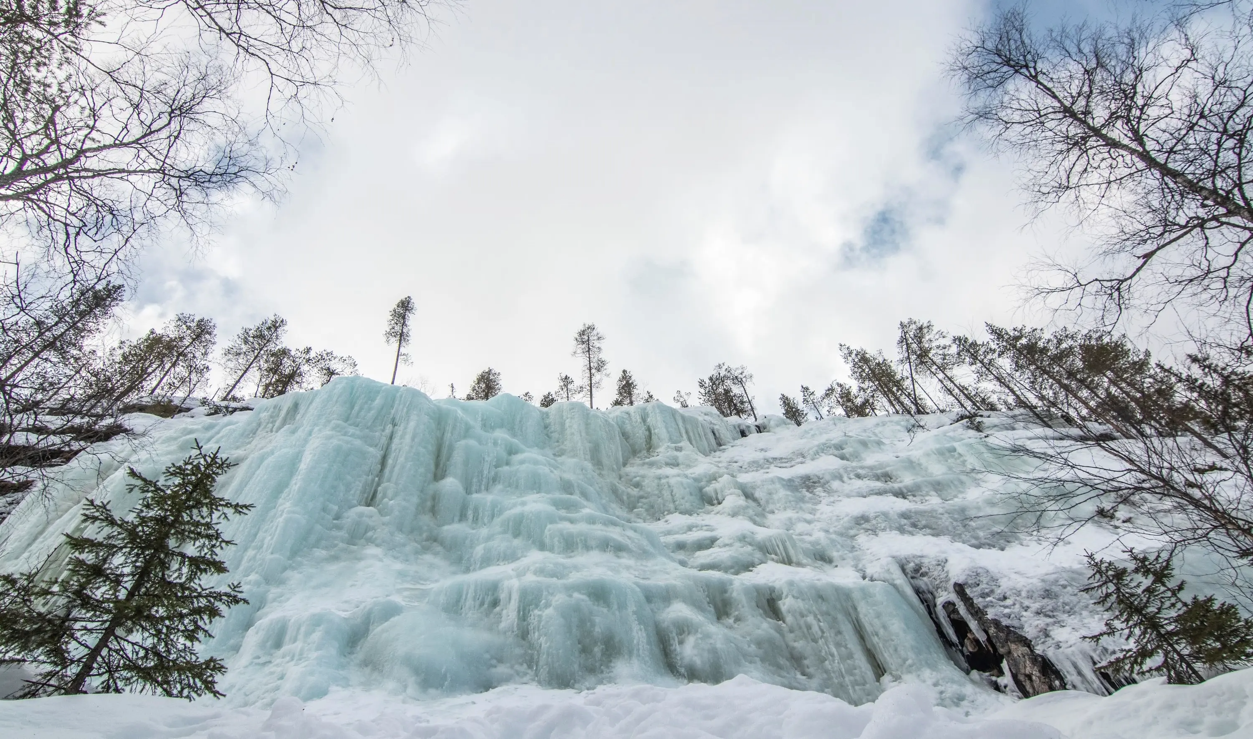 Frozen waterfall with ice formations surrounded by snow, with leafless bare trees and a partly cloudy sky in the background.